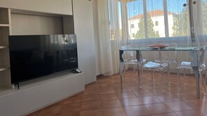 A modern living room with a flat-screen TV, clear glass dining table, and chairs.