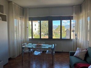 Uma área de refeições com uma mesa e cadeiras, uma janela com vista para prédios e árvores, e um sofá verde com um cobertor vermelho.