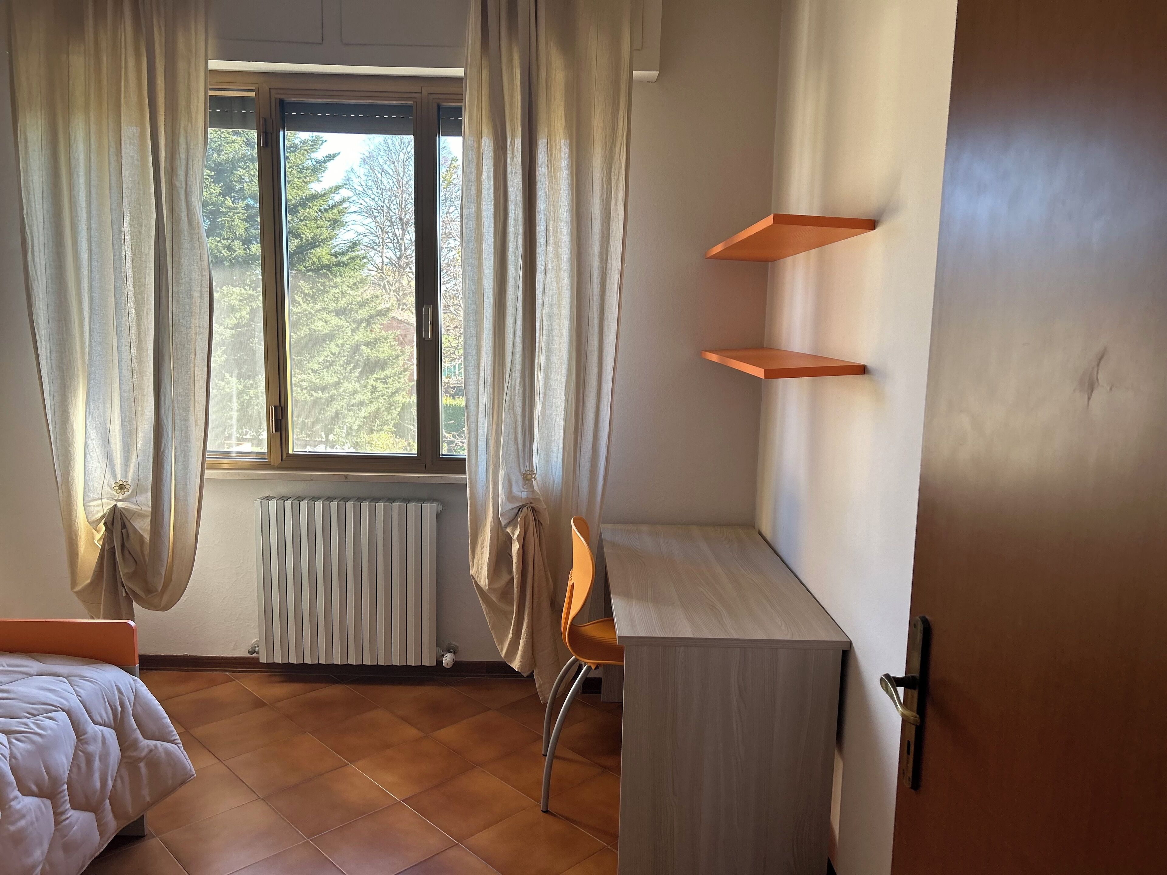 Une chambre à coucher avec un lit, un bureau et une chaise. Il y a une fenêtre avec des rideaux, un radiateur et une vue sur les arbres à l’extérieur.
