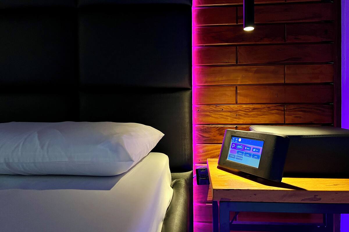 A hotel room with a bed, a bedside table with a digital device, and a brick-patterned wall.