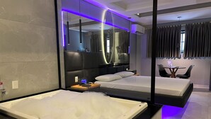 Una habitación de hotel moderna con una bañera grande, cama separada, una pequeña mesa con sillas y un espejo.