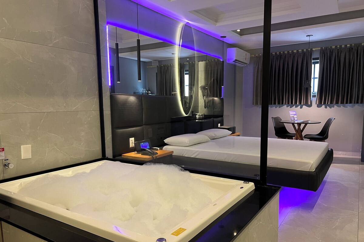 A modern hotel room with a large bathtub, a separate bed, a small table with chairs, and a mirror.