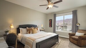Une chambre avec un grand lit, un bureau, une chaise, un ventilateur de plafond et une fenêtre donnant sur les montagnes.