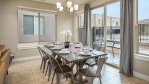 Un coin repas avec une table dressée pour six personnes, entourée de chaises, d’un lustre et de grandes fenêtres ornées de rideaux.