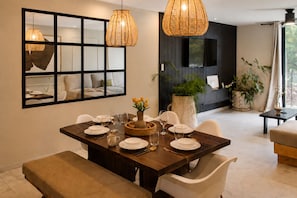 A dining area with a wooden table set for four, surrounded by white chairs. There are two large woven pendant lights above the table, and a large mirror on the wall.