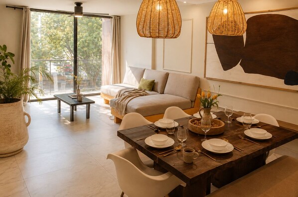 A dining area with a wooden table set for two, a sofa, and a large window with a view of trees and a red vehicle outside.