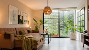 A modern living room with a sofa, a side table with a vase and a drink, a large window with a view of greenery, and a hanging light fixture. - Urban Stylish 3 BedRoom / Daily cleaning (Guadalajara)
