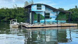 A floating house with a covered deck, a small boat docked nearby, and a mural depicting a natural landscape. - The Burrow, an amazing floating cabin with pontoon rental available (Grant-Valkaria)