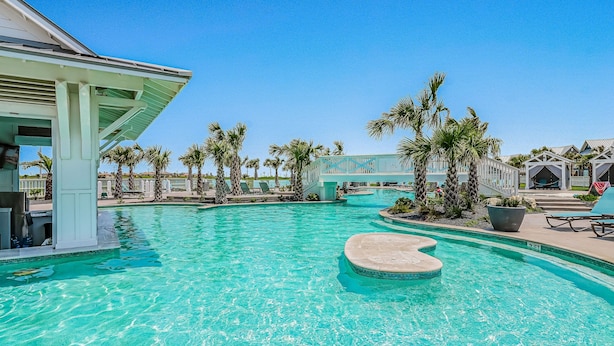 A swimming pool with a heart-shaped island, surrounded by palm trees and lounge chairs. - Gorgeous family beach home with balcony and wonderful amenities (Port Aransas)