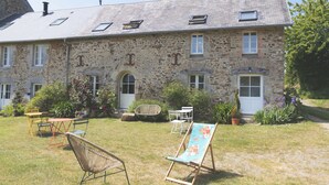 A stone building with a sloped roof, multiple windows, and a small garden with outdoor furniture. - ESCALE DE GERBOLD (Gratot)