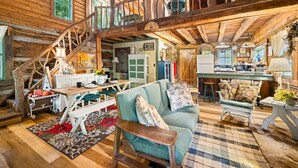 A rustic cabin interior with a wooden staircase, a dining area with a table and chairs, a kitchen with a refrigerator, and a living room with a sofa and a coffee table.