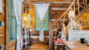A rustic cabin interior with wooden walls, a staircase, a dining table, and a window with curtains.