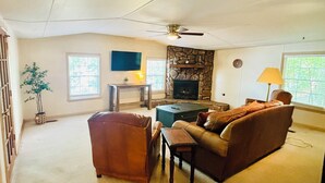 A spacious living room with a stone fireplace, a ceiling fan, and a television mounted on the wall.