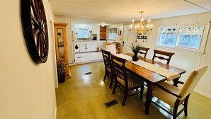 A dining area with a wooden table and chairs, a chandelier, and a kitchen in the background.