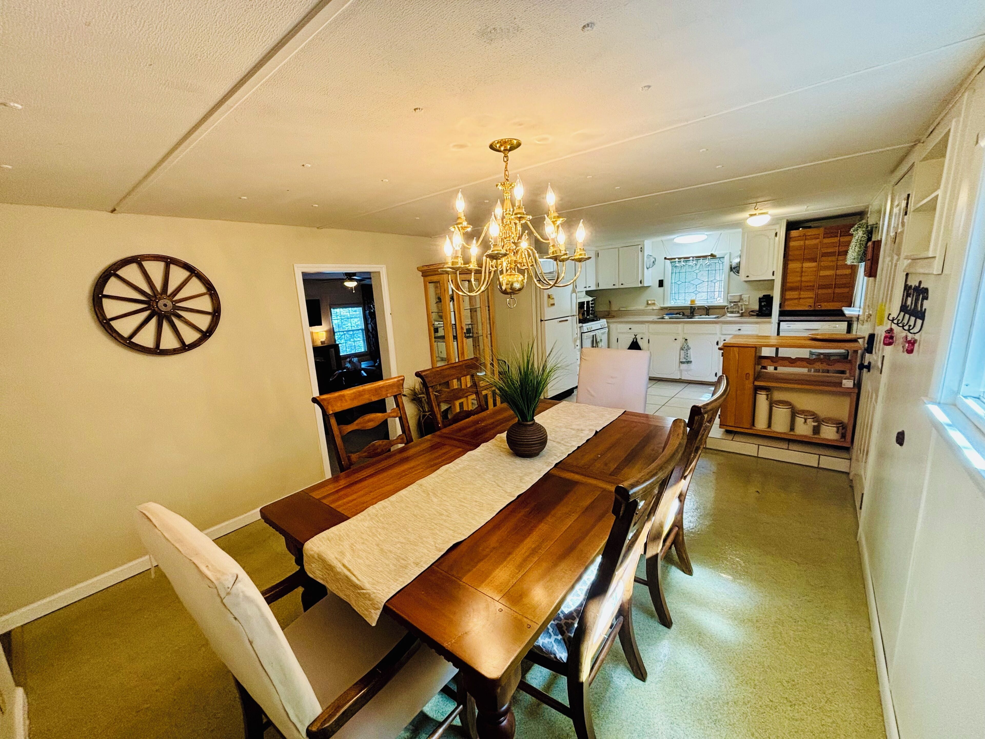 A dining area with a wooden table, chairs, and a chandelier. There is a kitchen in the background with white cabinets and a window.