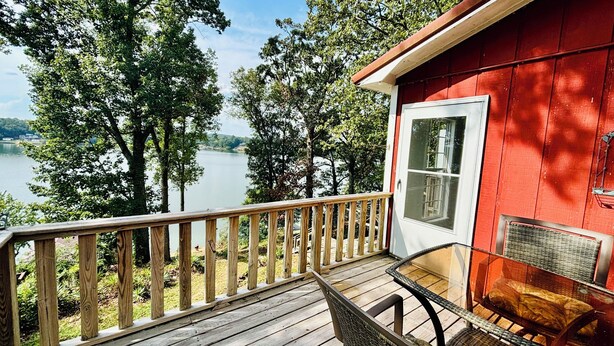 A wooden deck with a glass table and chairs overlooking a lake, a red building with a white door, and a metal grill attached to the building.
