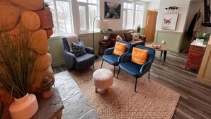 A living room with a stone fireplace, two blue armchairs with orange cushions, a round ottoman, and a large window with a view of trees. - Three Peaks Getaway 2 bedrooms & 2 bath-sleeps 6 (Canmore)