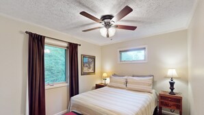 Ein Schlafzimmer mit Bett, Deckenventilator, Fenster mit Vorhängen, Nachttisch mit Lampe und einem kleinen Teppich mit Bär-Motiv.