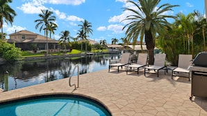 A pool area with lounge chairs, a grill, and a view of a house and palm trees.