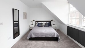 A bedroom with a sloped ceiling, a bed with a grey quilt, two bedside tables with lamps, a large mirror, and a radiator. - Hampden Apartments - The William (Windsor)