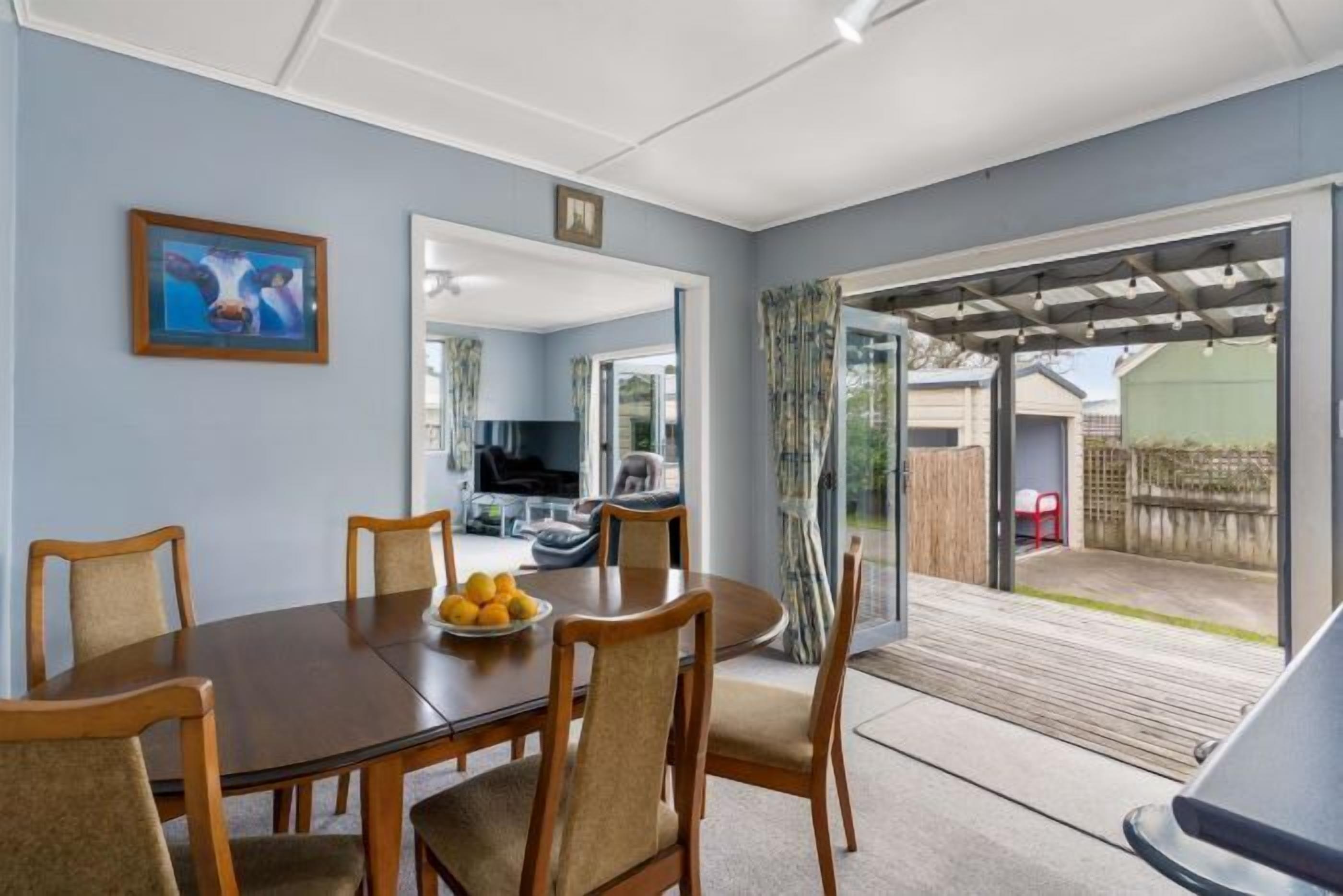 A dining area with a round table, chairs, and a bowl of fruit. There is a large window and a door leading to a patio.