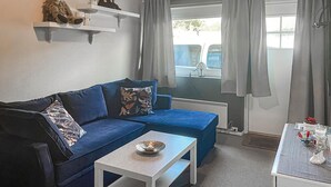 Una habitación acogedora con un sofá azul, una mesita blanca con un cuenco, y un radiador. Hay estantes con objetos decorativos y una ventana con cortinas.