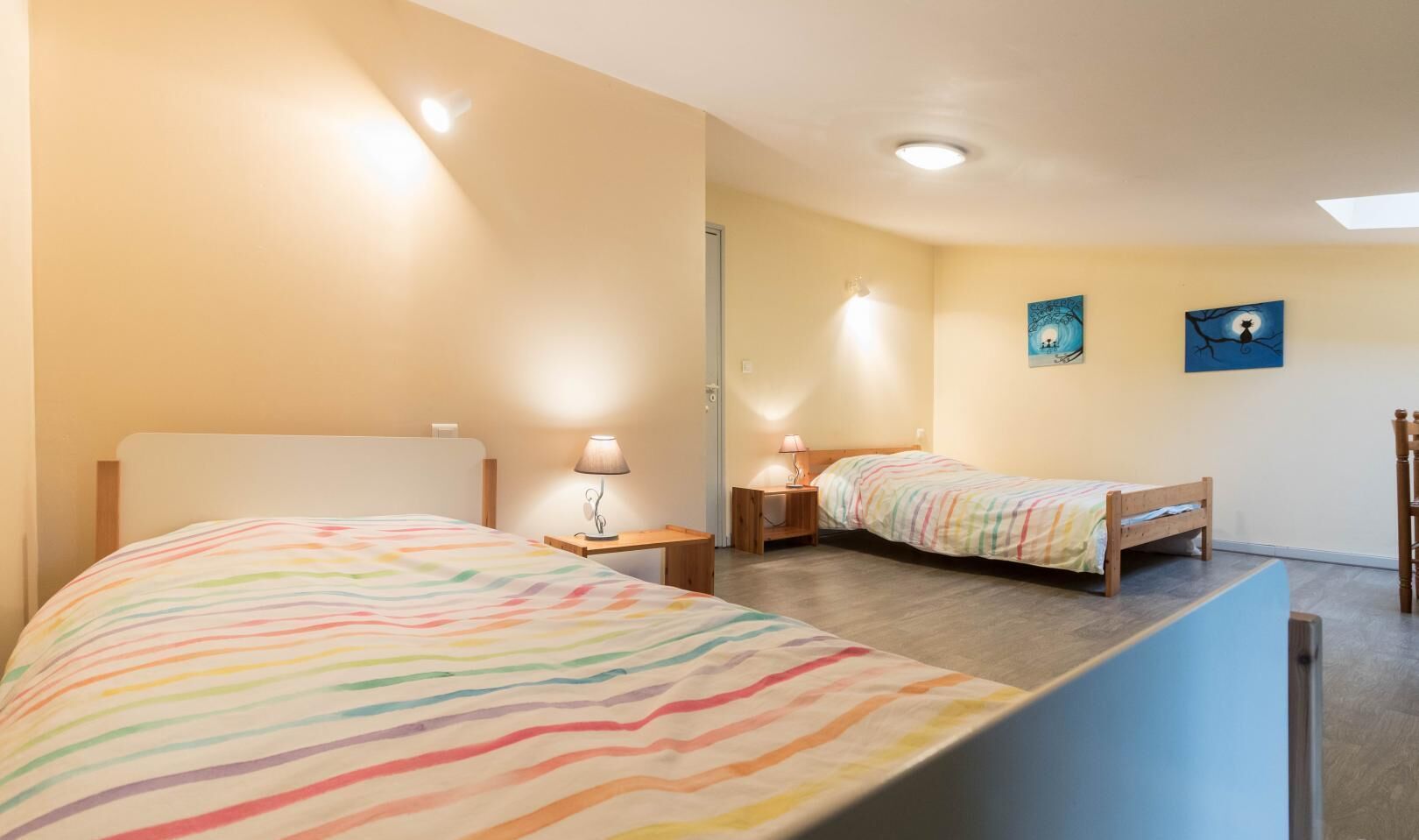 Habitación de hotel con dos camas, cada una con una colcha a rayas de colores, una mesita de noche con lámpara y un cuadro colgado en la pared.