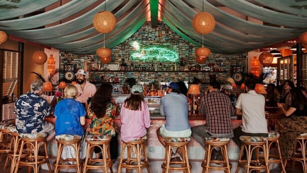Un bar con clientes sentados en taburetes, un letrero de neón y una estantería llena de libros detrás de la barra.
