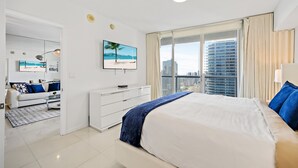 A modern hotel room with a large bed, a flat-screen TV, a city view, and a minimalist decor.