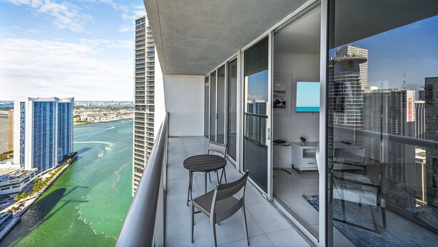 A balcony with a view of a river and cityscape, featuring a table and chairs, a small plant, and a glass partition.