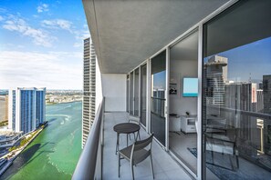En balkon med udsigt over en flod og bybillede, med et bord og stole, en lille plante og et glasgelænder.