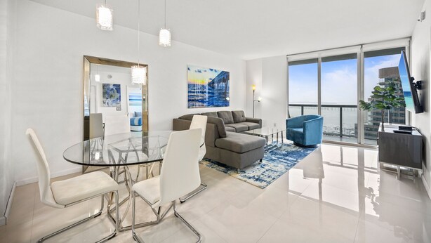 Un salon moderne avec une table à manger en verre, des chaises blanches, un canapé gris et un fauteuil bleu. Une grande fenêtre offre une vue sur l’océan et un immeuble.