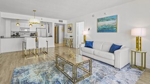 A modern living room with a glass coffee table, white sofa, and blue accent pillows. There is a kitchen in the background with white cabinets and gold fixtures. - The Club 3904 · High Floor Unit Located in Brickell - Free Parking (Miami)