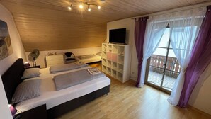 Ein Schlafzimmer mit Holzboden, einem Bett mit karierter Bettwäsche, einem Fernseher auf einer weißen Kommode und einem Balkon mit lila Vorhängen.