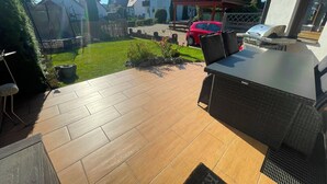 Eine Terrasse mit Tisch und Stühlen, im Hintergrund ein rotes Auto, und ein Haus mit weißer Fassade.