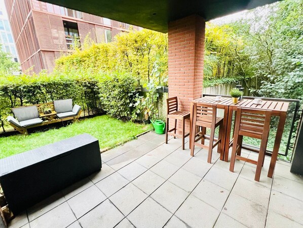 A patio with a wooden table and chairs, a black sofa, and a small garden area with a hedge and a brick wall.