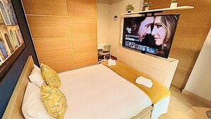 A modern hotel room with a large bed, a flat-screen TV mounted on the wall, a small desk, and a shelf with decorative items. - Chic one bedroom with parking&terrace (Luxembourg City)