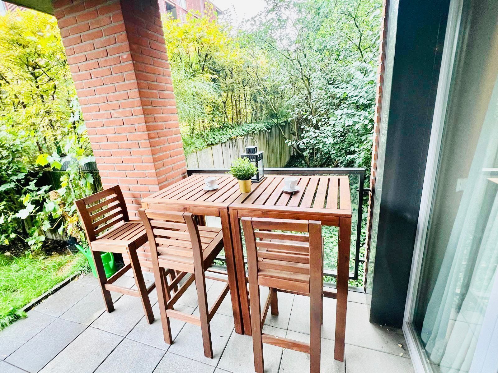 Une table et des chaises en bois sur un balcon, avec un mur en briques et de la verdure en arrière-plan.