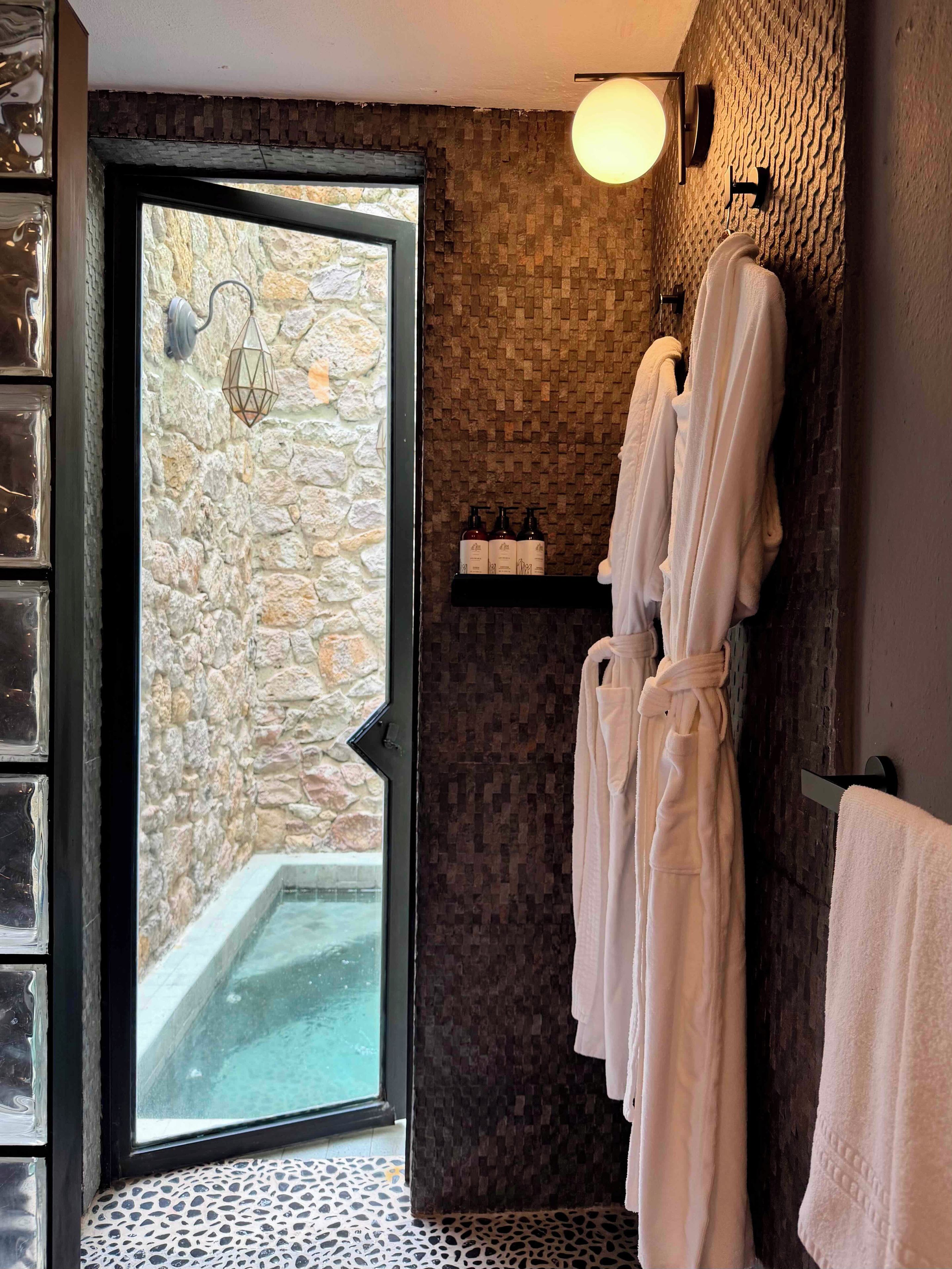 Un baño con pared de piedra, una puerta de cristal que da al exterior y un perchero con toallas blancas.
