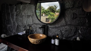 A bathroom with a stone wall, a round mirror, a wooden basin, and two bottles of lotion. - Casa Nueve (San Miguel de Allende)