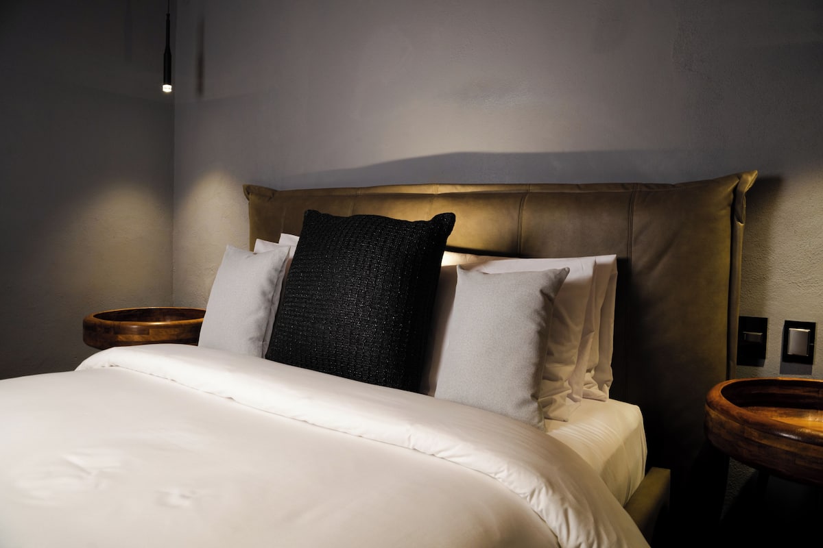 Una cama bien hecha con sábanas blancas, un cojín de textura oscura y una mesita de noche de madera con lámpara.