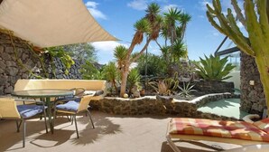 An outdoor patio with a round table and chairs, a lounge chair, and a variety of cacti and palm plants. - CASA VERDE Apartment 4 Detached small house. Sea view Risco Famara (CN)