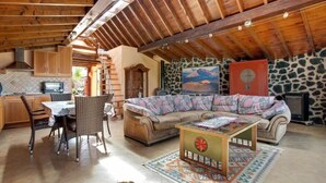 A cozy living area with a sofa, coffee table, and dining table. There is a kitchen area with cabinets and a microwave. The ceiling is wooden, and there is a stone wall with a painting. - CASA VERDE Apartment 4 Detached small house. Sea view Risco Famara (CN)