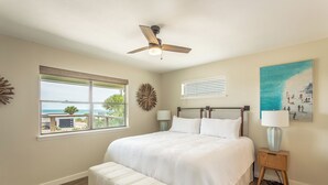 Una camera da letto con un letto, una ventola a soffitto, una finestra vista oceano, un quadro e un comodino.