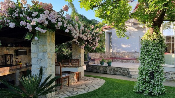 A stone patio with a dining area, a stone wall with climbing roses, and a white building with large windows.