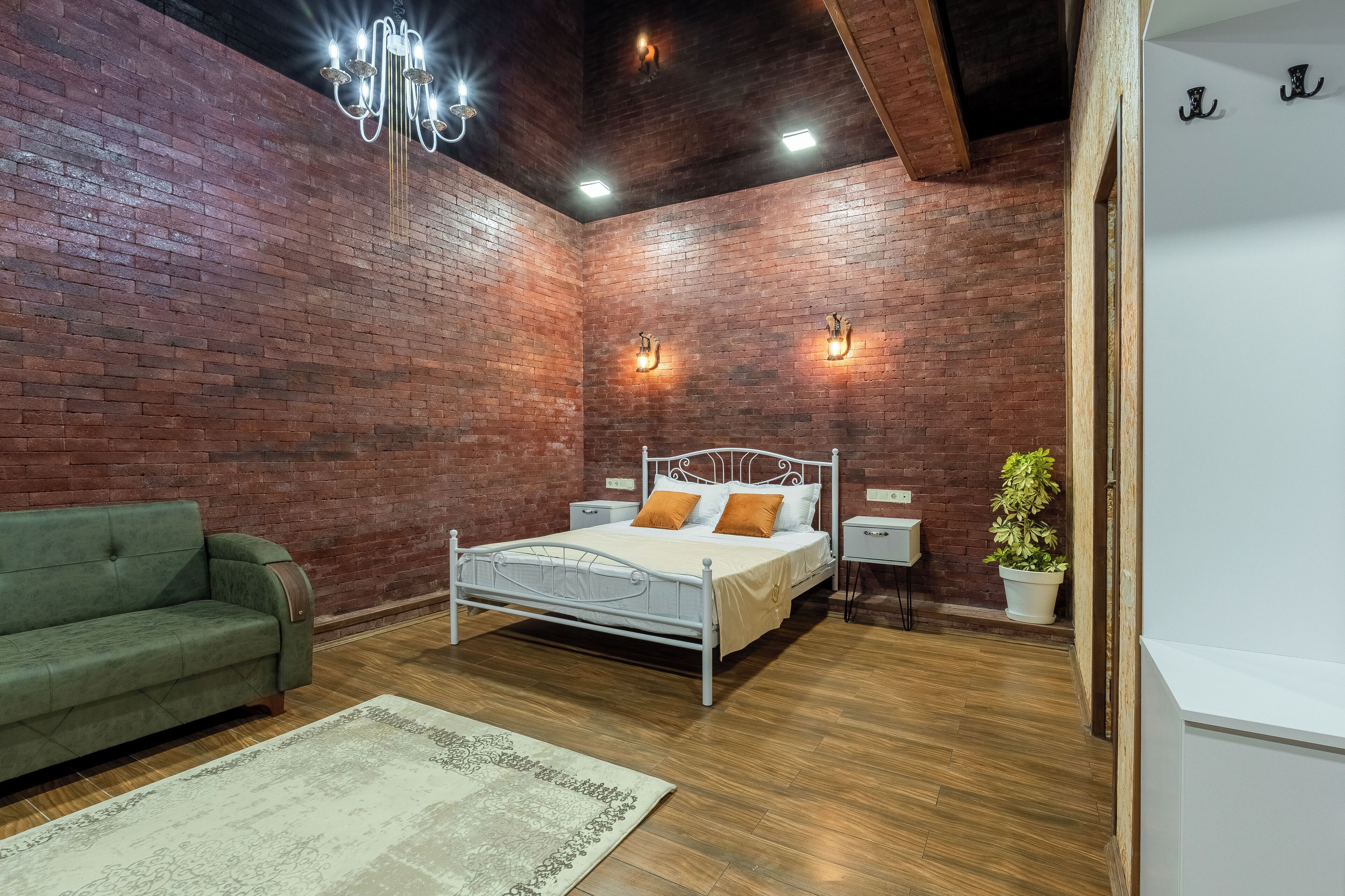 Ein Schlafzimmer mit Backsteinwand, einem grünen Sofa, einem weißen Bett mit orangenen Kissen, Holzboden und einer Topfpflanze.