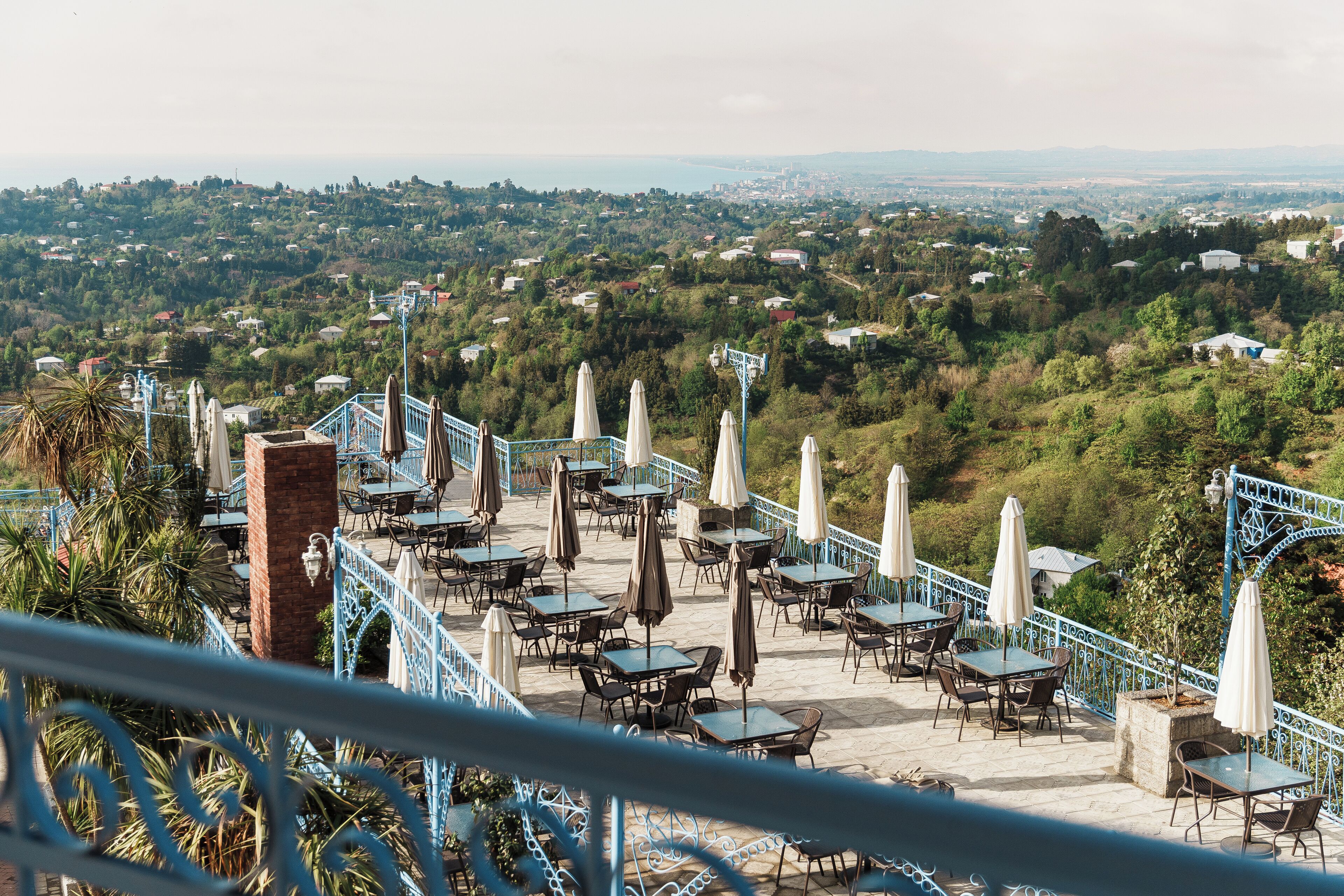 An outdoor terrace with tables and chairs, surrounded by a blue railing, overlooking a hilly landscape with buildings.