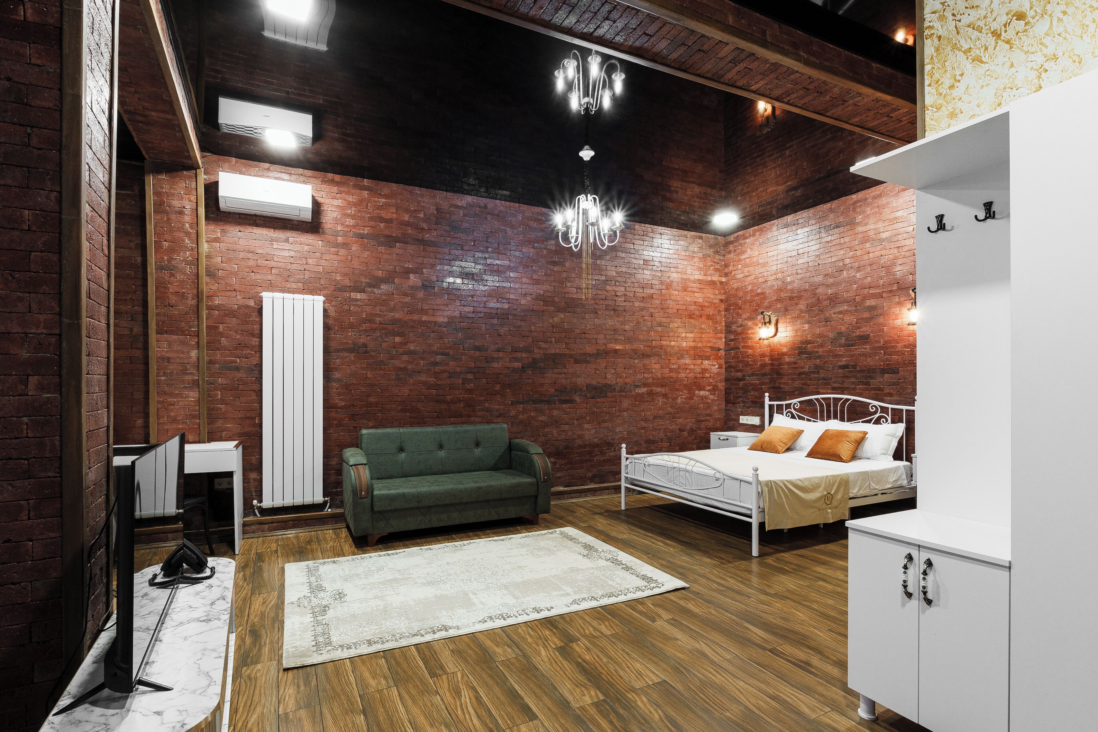 A room with a brick wall, a bed with white bedding, a green sofa, a white cabinet, and a chandelier.
