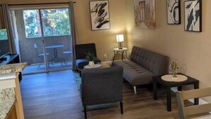Ein modernes Wohnzimmer mit einer Couch, einem Sessel und einem Esstisch. An den Wänden befinden sich abstrakte Gemälde und eine Glastür führt in einen Außenbereich.