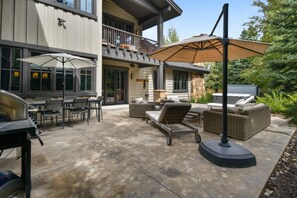 Een terras buiten met een barbecue, eettafels, stoelen, een bank en een grote parasol.